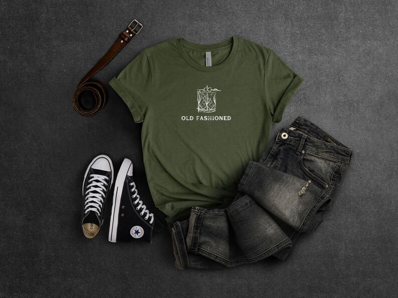 May include: A green t-shirt with a white graphic of a cocktail glass and the text "Old Fashioned". The t-shirt is laid out on a gray surface with a pair of black sneakers and a brown leather belt.