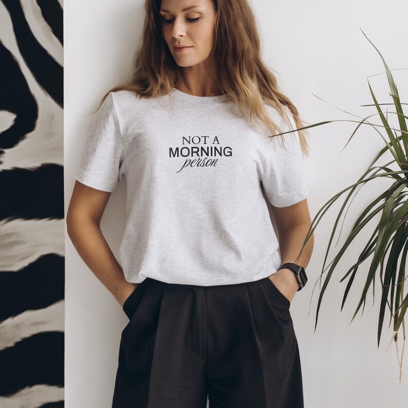 Pu&ograve; includere: Una donna indossa una maglietta grigia con la scritta "NOT A MORNING person" stampata su di essa. Indossa pantaloncini neri e ha le mani in tasca.