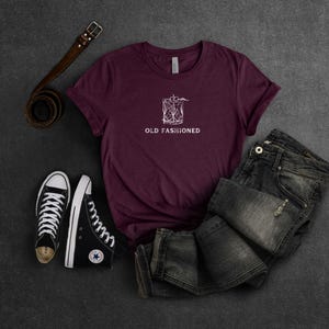 May include: A maroon t-shirt with a white graphic of a cocktail glass and the text "Old Fashioned". The shirt is laid out on a dark surface with a pair of black high-top sneakers and a brown leather belt.
