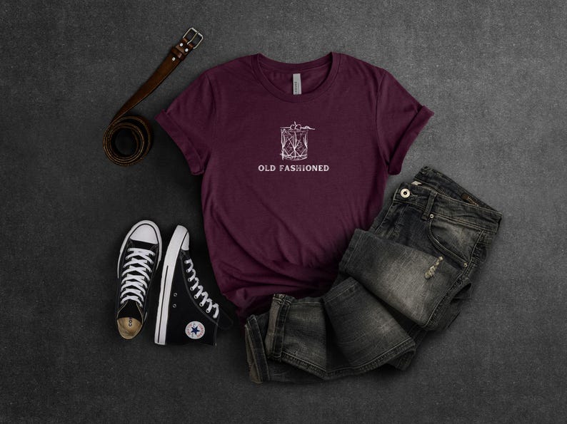 May include: A maroon t-shirt with a white graphic of a cocktail glass and the text "Old Fashioned". The shirt is laid out on a dark surface with a pair of black high-top sneakers and a brown leather belt.