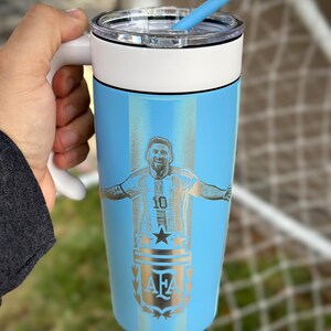 May include: A light blue insulated travel mug with a white handle and lid. The mug has a silver engraving of a soccer player with the number 10 and the letters AFA. A blue straw is inserted into the lid.