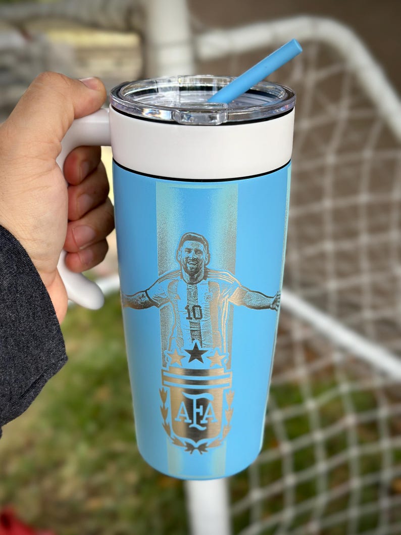 May include: A light blue insulated travel mug with a white handle and lid. The mug has a silver engraving of a soccer player with the number 10 and the letters AFA. A blue straw is inserted into the lid.