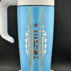 May include: A light blue travel mug with a white handle and lid. The mug features a silver emblem with the letters "AFA" and two stars, and a silver stripe design. The mug is approximately 18 cm tall.
