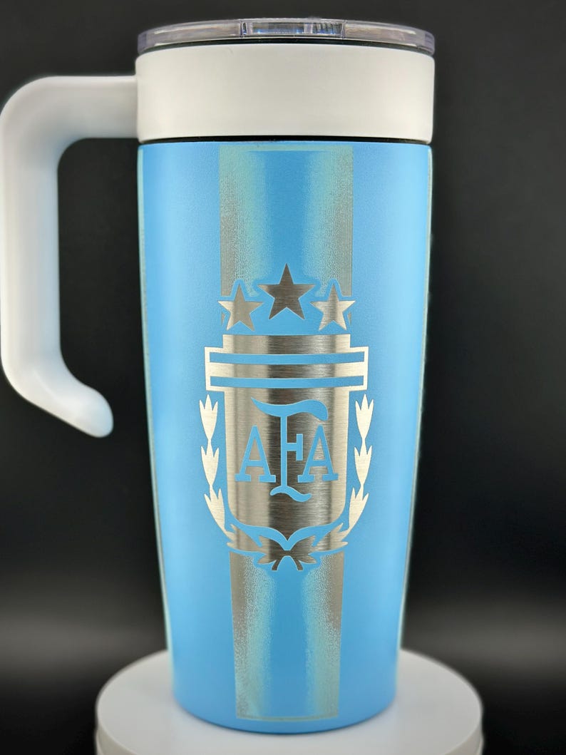 May include: A light blue travel mug with a white handle and lid. The mug features a silver emblem with the letters "AFA" and two stars, and a silver stripe design. The mug is approximately 18 cm tall.