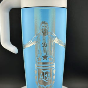 May include: A light blue travel mug with a white handle and lid. The mug features a silver engraving of a soccer player in a jersey with the number 10, along with the AFA logo and stars.