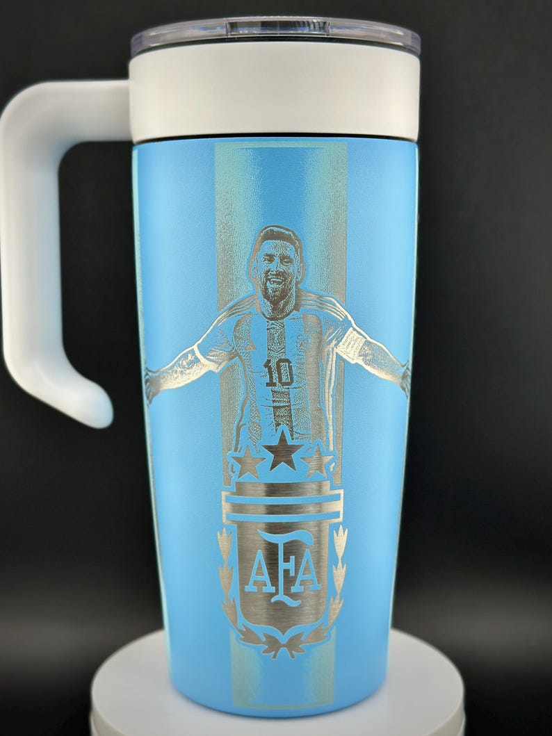 May include: A light blue travel mug with a white handle and lid. The mug features a silver engraving of a soccer player in a jersey with the number 10, along with the AFA logo and stars.