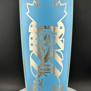 May include: A light blue insulated tumbler with a silver design. The design includes a maple leaf, stars, and a trophy. The tumbler has a clear lid and is cylindrical in shape.