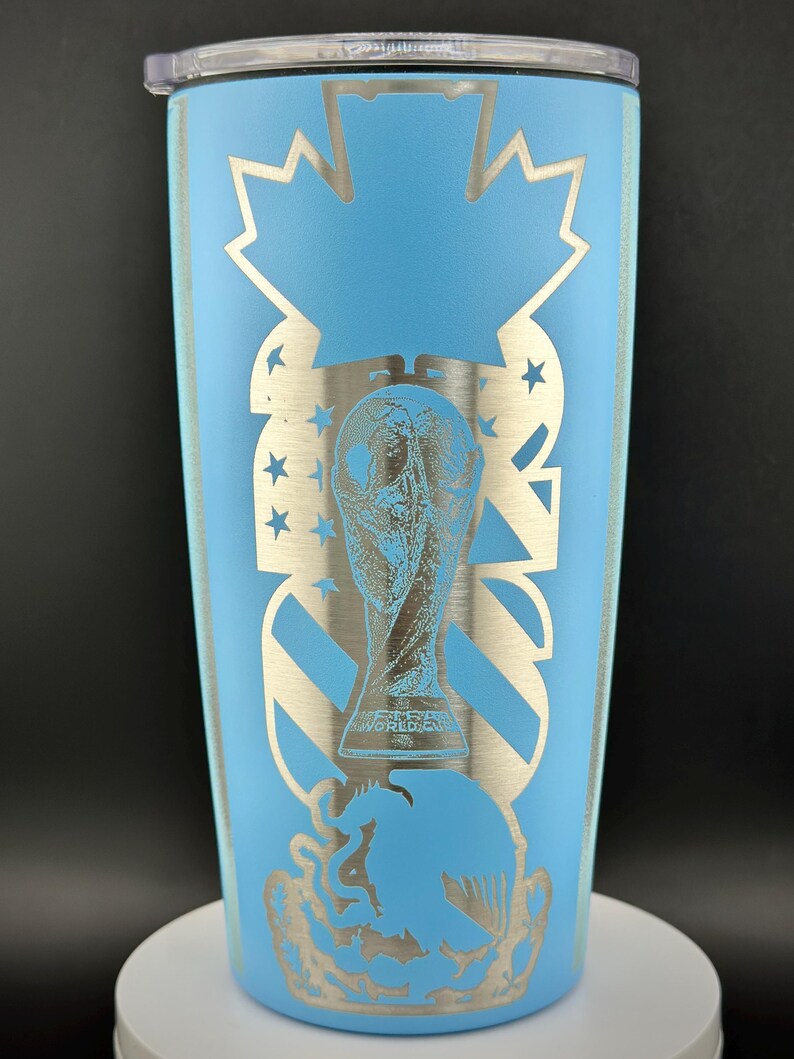 May include: A light blue insulated tumbler with a silver design. The design includes a maple leaf, stars, and a trophy. The tumbler has a clear lid and is cylindrical in shape.