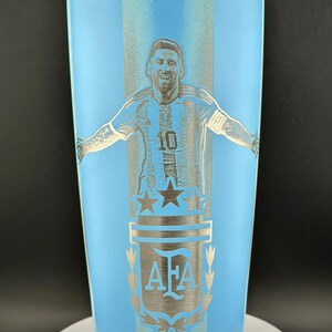 May include: A light blue insulated tumbler featuring a silver engraving of a soccer player with arms outstretched. The design includes the number 10, stars, and the letters "AFA". The tumbler has a black lid and is approximately 20 cm tall.