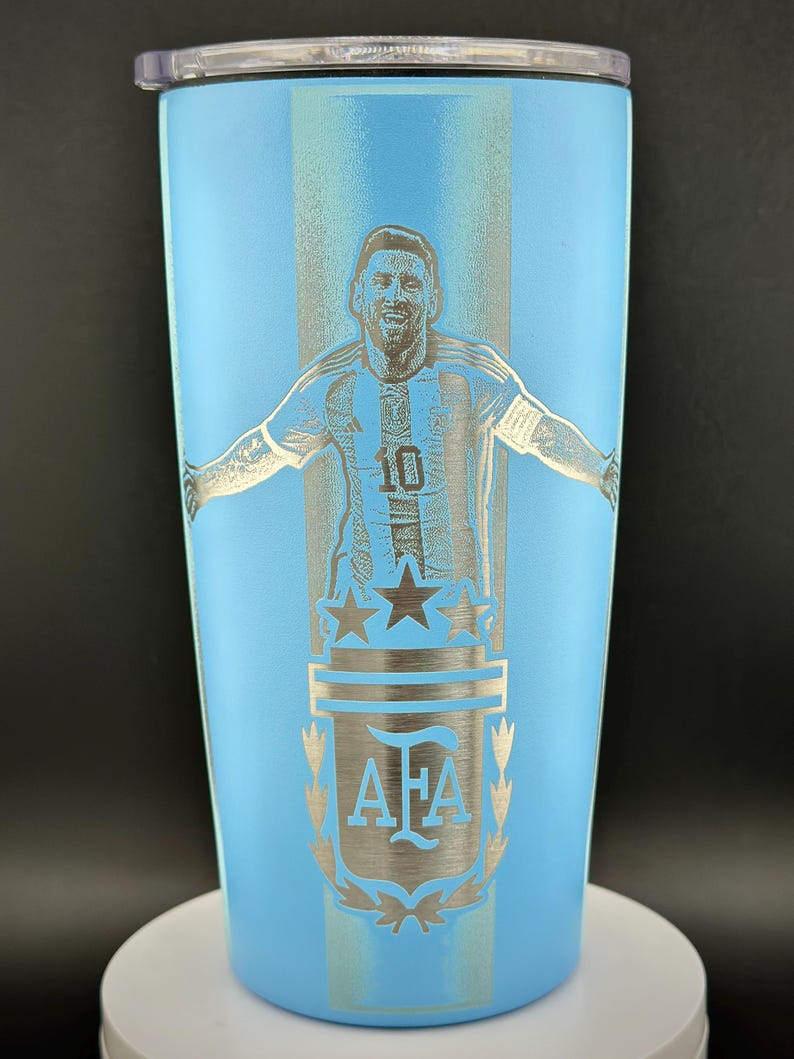 May include: A light blue insulated tumbler featuring a silver engraving of a soccer player with arms outstretched. The design includes the number 10, stars, and the letters "AFA". The tumbler has a black lid and is approximately 20 cm tall.