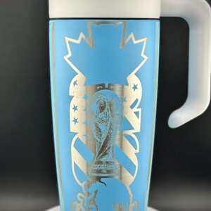 May include: A light blue travel mug with a white handle and lid. The mug features a silver design of the World Cup trophy, stars, and a maple leaf. The mug is approximately 18 cm tall.