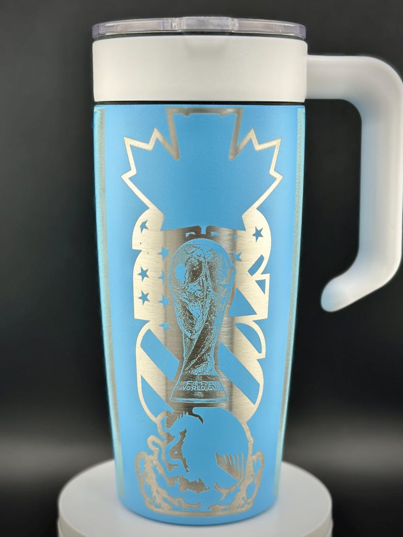 May include: A light blue travel mug with a white handle and lid. The mug features a silver design of the World Cup trophy, stars, and a maple leaf. The mug is approximately 18 cm tall.