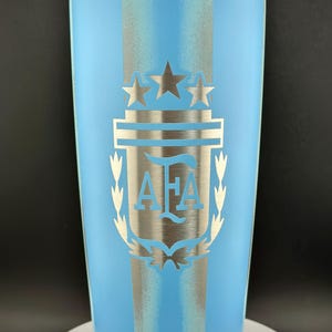 May include: A light blue insulated travel mug with a silver emblem. The emblem features three stars, a shield, and the letters "AFA". The mug has a silver stripe down the centre and a clear lid. The design is likely related to a sports team or organization.