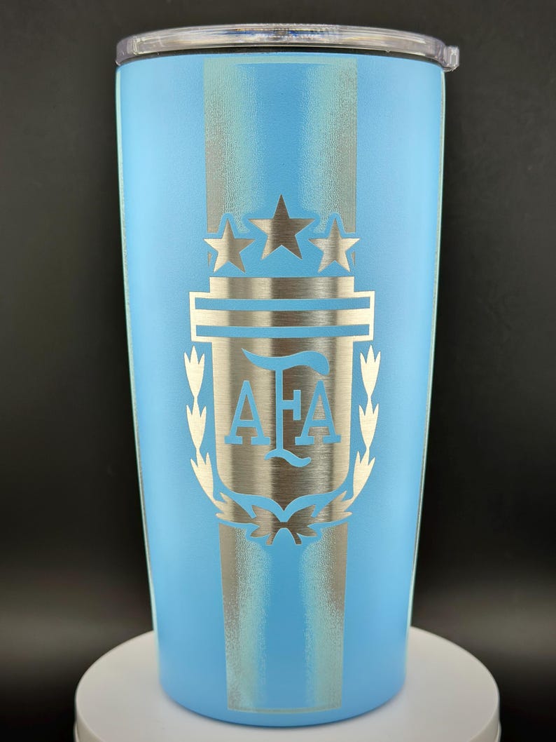 May include: A light blue insulated travel mug with a silver emblem. The emblem features three stars, a shield, and the letters "AFA". The mug has a silver stripe down the centre and a clear lid. The design is likely related to a sports team or organization.