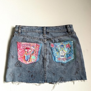 May include: A denim skirt with two painted pockets. The left pocket features a pink alien with the text "EMERGING" and the right pocket features a blue alien with the text "INTERCOM".