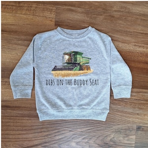 May include: A light gray sweatshirt featuring a green combine harvester graphic with wheat. The text "Dibs on the Buddy Seat" is printed below the image. The long-sleeved crew neck shirt is made of a soft material.