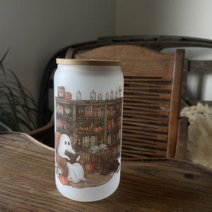 May include: A white frosted glass jar with a bamboo lid features a whimsical illustration of a ghost reading a book in a cozy library setting. The scene includes bookshelves filled with books, candles, pumpkins, and a black cat.