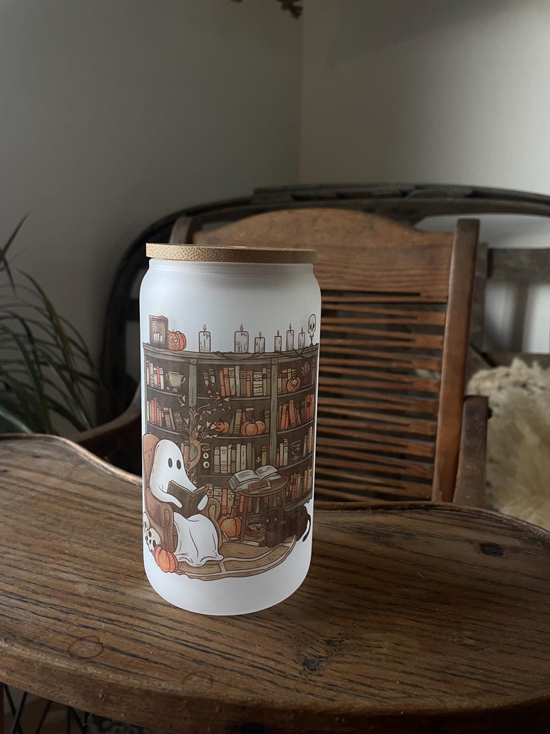 May include: A white frosted glass jar with a bamboo lid features a whimsical illustration of a ghost reading a book in a cozy library setting. The scene includes bookshelves filled with books, candles, pumpkins, and a black cat.