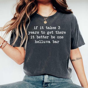 May include: Dark grey t-shirt with the text "if it takes 3 years to get there it better be one helluva bar" in white. The shirt is a casual wear item.