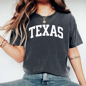 May include: A charcoal gray short-sleeve t-shirt featuring the word TEXAS printed in large, bold, white capital letters across the chest in a classic collegiate-style arched font. The garment has a crew neckline and a relaxed, casual fit.