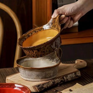 May include: Two ceramic serving bowls, one filled with a golden liquid, stacked on a wooden cutting board. The top bowl has a handle and a speckled brown glaze. The bottom bowl is gray and white.