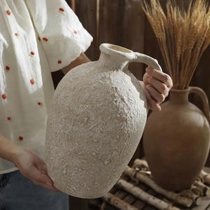 Puede incluir: Una jarra grande de cerámica blanquecina texturizada con asa. La jarra la sostiene una persona. En el fondo, hay una jarra marrón y un manojo de espigas de trigo. La jarra tiene un aspecto rústico y envejecido.