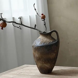 Op de afbeelding: Een bruine keramische vaas met een handvat, met een textuur en donkere accenten. Een tak met kleine, rood-oranje vruchten steekt uit de vaas. De vaas staat op een houten oppervlak.