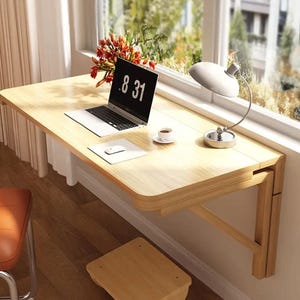 Puede incluir: Un escritorio de madera clara montado en la pared con una computadora portátil, un ratón y una lámpara. La pantalla de la computadora portátil muestra "8 31". Una taza pequeña de café y una planta también están en el escritorio. El escritorio está soportado por soportes de madera.