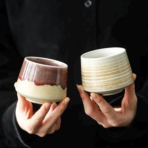 May include: Two ceramic cups are held in hands against a dark background. One cup is a gradient of burgundy and cream, the other is cream with horizontal gold stripes. Both have a unique, angled design.