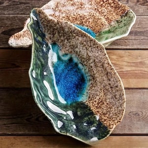 Puede incluir: Dos fuentes decorativas de cerámica con formas orgánicas. Las fuentes presentan una superficie texturizada y moteada en tonos marrones, verdes y azules. El interior de una fuente tiene un centro azul vibrante, mientras que la otra tiene un borde verde.