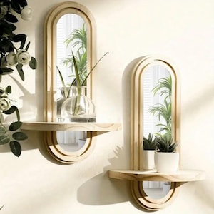Puede incluir: Dos espejos de pared de madera con marcos arqueados y estantes integrados. Cada espejo refleja vegetación y está adornado con un estante debajo, con un espejo semicircular. Un estante sostiene un jarrón de vidrio, el otro plantas en macetas.