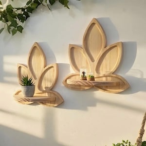 May include: Two wooden lotus flower-shaped wall shelves. Each shelf has a small, flat shelf area. One shelf holds a small potted plant, and the other holds two miniature glass terrariums. The shelves are made of light-colored wood.