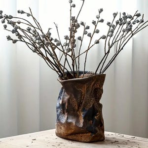 Puede incluir: Un jarrón de cerámica marrón con diseño de bolsa de papel arrugada, lleno de ramas secas con pequeñas cápsulas de semillas grises. El jarrón tiene una superficie texturizada y está sobre una mesa de madera, creando una estética natural y terrosa.