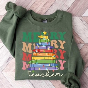 May include: A dark green sweatshirt with the words "MERRY teacher" in a colorful, stacked design resembling a Christmas tree made of books. The design includes a star and string lights.