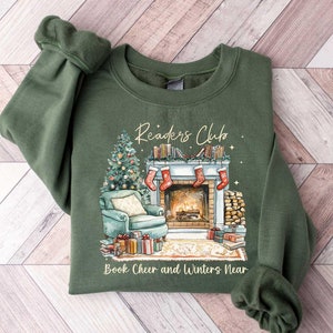 K&ouml;nnte beinhalten: Ein gr&uuml;nes Sweatshirt mit einem weihnachtlichen Design, das einen Kamin, einen Weihnachtsbaum und B&uuml;cher zeigt. Der Text "Readers Club" befindet sich &uuml;ber dem Kamin und "Book Cheer and Winters Near" unter dem Kamin.
