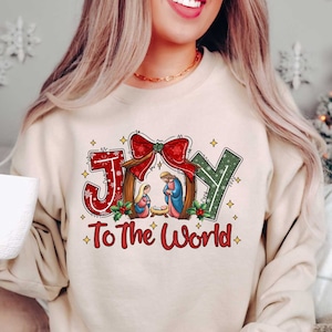 May include: A beige sweatshirt with a Christmas-themed design featuring the word "JOY" with a red bow and a nativity scene. The text "To The World" is printed below the word "JOY".