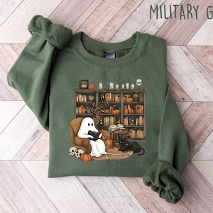 May include: A green sweatshirt with a graphic of a ghost reading a book in a chair next to a bookcase and a black cat. The text "MILITARY GREEN" is in the upper right corner of the image.