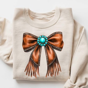 May include: A beige sweatshirt with a brown leather bow with turquoise gemstone embellishments and fringe details. The word "SAND" is printed in brown text in the upper right corner of the image.
