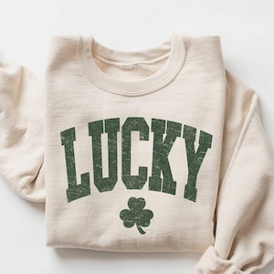 Może przedstawiać: Beżowa bluza z zieloną koniczyną i nadrukiem słowa "LUCKY" w stylu vintage.