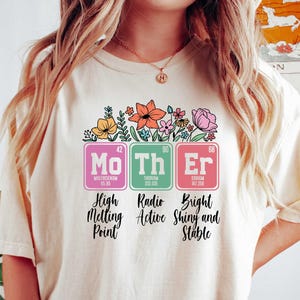 May include: Cream-colored t-shirt with a floral design above the word "Mother" spelled out using periodic table elements. The shirt features the words "High Melting Point", "Radio Active", and "Bright Shiny and Stable" below the elements.