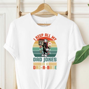 May include: White t-shirt with a retro-style graphic of a cassette tape with the text "I keep all my dad jokes in a dad-a-base".