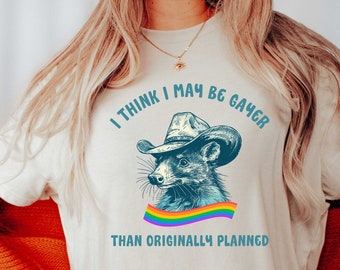 May include: A white t-shirt with a cartoon raccoon wearing a cowboy hat and a rainbow ribbon. The text on the shirt reads "I think I may be gayer than originally planned."