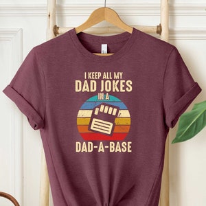 May include: A maroon t-shirt with a retro-style graphic that says "I keep all my dad jokes in a dad-a-base". The graphic features a floppy disk with a sunset background.