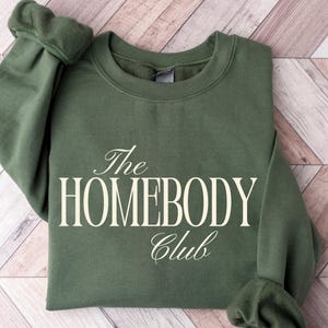 May include: An olive green sweatshirt with the text "The HOMEBODY Club" in a white, elegant font. The sweatshirt is folded neatly on a light brown and white wooden surface. The sleeves are cuffed.