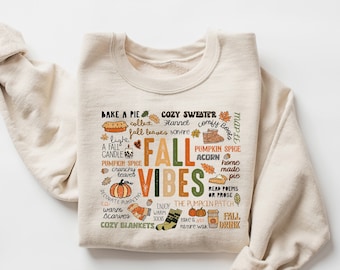 May include: A beige sweatshirt with a graphic print of fall-themed words and illustrations. The text includes "Fall Vibes", "Bake a Pie", "Cozy Sweater", "Pumpkin Spice", "Home Made Pie", and "The Pumpkin Patch".