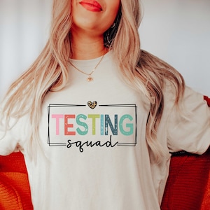 May include: A white oversized t-shirt with a colorful graphic that says "TESTING squad" with a leopard print heart above the text.