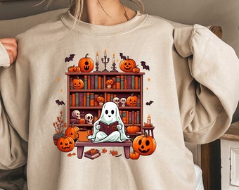 May include: Beige sweatshirt featuring a Halloween-themed graphic. The design includes a ghost reading a book in front of a bookshelf with pumpkins, candles, and bats. The sweatshirt is a comfortable, casual style.
