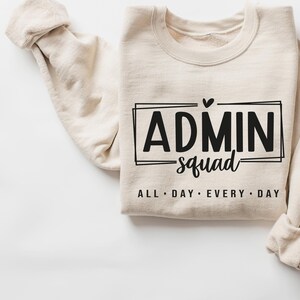 May include: A beige sweatshirt with the text "ADMIN squad ALL DAY EVERY DAY" printed on the front. The text is in a black font and is surrounded by a black rectangle.