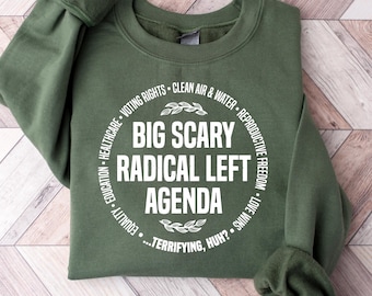 Op de afbeelding: Een olijfgroene sweatshirt met de tekst "BIG SCARY RADICAL LEFT AGENDA" in een wit cirkelvormig ontwerp. Het ontwerp bevat zinnen als "Stemrecht" en "Reproductieve Vrijheid". De sweatshirt wordt gepresenteerd op een licht houten oppervlak.