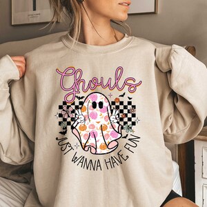 May include: A beige sweatshirt with a pink and white ghost graphic. The ghost is patterned with pink and orange pumpkins and flowers. The text "Ghouls Just Wanna Have Fun" is printed below the ghost.